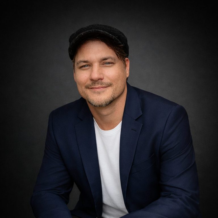 Mann in einem lässigen Anzug und einer Schirmmütze sitzt lächelnd vor dunklem Hintergrund.