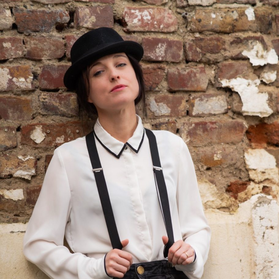 Frau in schwarzem Hut und Hosenträgern vor einer gemusterten Backsteinwand.